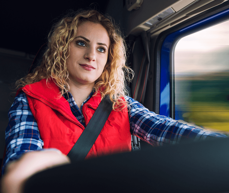 imagem de caminhoneira dirigindo. Indicando a inclusão de mulheres como motoristas de caminhão. Mulher de pele clara, cabelos loiros cacheados, vestindo camisa xadrez azul de mangas compridas e colete vermelho.